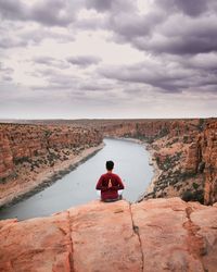 Rear view of man sitting on cliff against sky