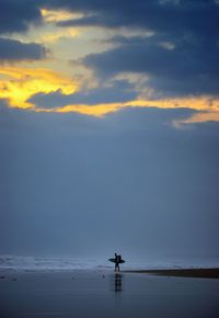 Distant view of man with surfboard walking at beach during sunset