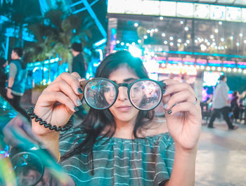 Woman holding eyeglasses while standing against illuminated lights