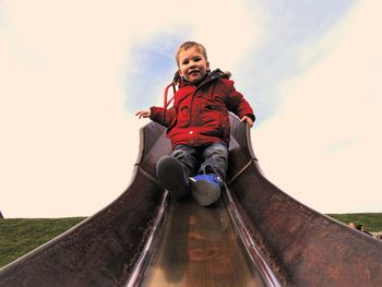 Low angle portrait of boy sliding on slide against cloudy sky