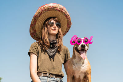 Low angle view of young woman wearing cowboy hat while standing with dog against clear sky