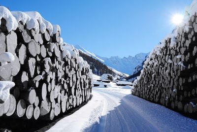 Road amidst stacked logs leading towards snowcapped mountain against sky