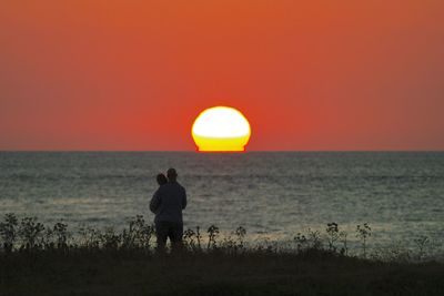 Rear view of couple walking at beach against sky during sunset