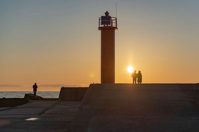 Silhouette people on lighthouse by sea against sky during sunset