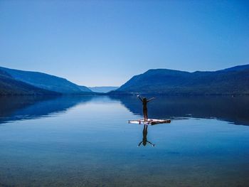 Woman with arms raised standing on wooden raft in lake against clear blue sky