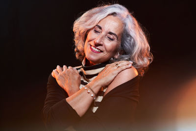 Happy senior female in stylish clothes with gray hair smiling with closed eyes and touching shoulders with crossed arms against black background