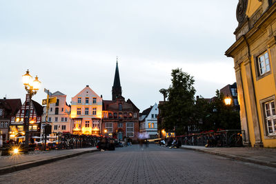 Illuminated street amidst buildings in city against sky at dusk