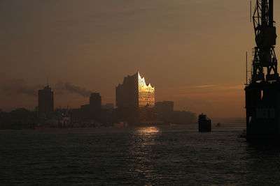 Silhouette buildings by sea against sky during sunset