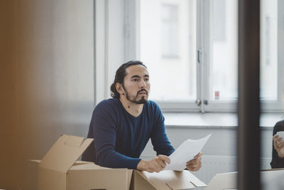 Young businessman holding document while leaning on cardboard boxes in new office