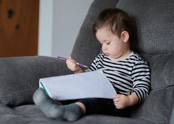 Young baby boy toddler writing in a notebook on the couch at home
