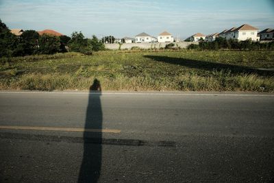 Shadow of person on road amidst field against buildings