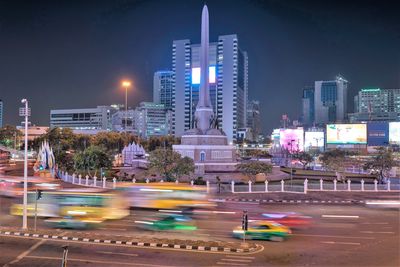 Light trails on road by buildings against sky at night