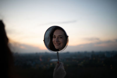 Reflection of smiling woman in mirror against sky during sunset