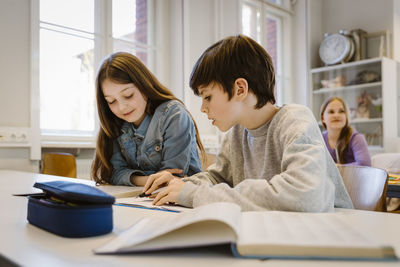 Boy helping female friend while studying together sitting at desk in classroom