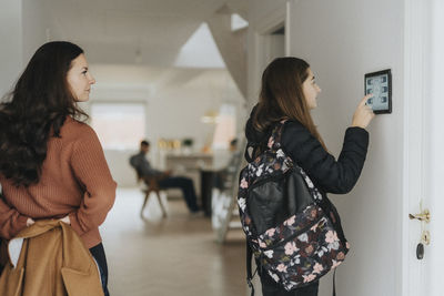 Girl with backpack using digital tablet mounted on wall by mother at home