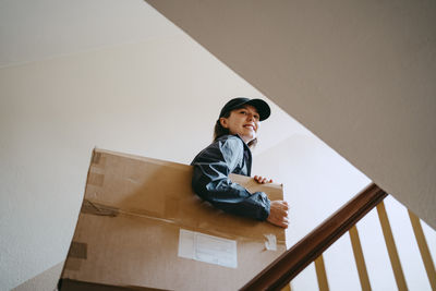 Low angle view of smiling female delivery person holding cardboard box while moving up on staircase in building