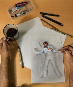 Cropped hand of young man holding coffee cup while drawing on paper at table