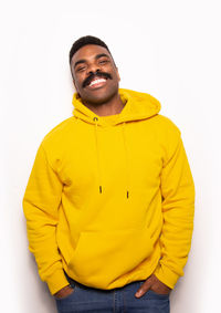 Positive african american man in bright yellow hoodie smiling and looking at camera while standing against white background