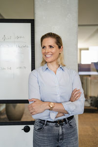 Smiling businesswoman with arms crossed leaning on column at workplace
