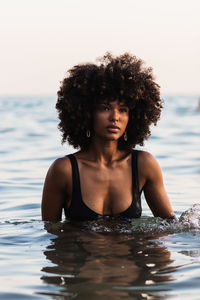 Tranquil black female wearing bikini standing in calm sea water in summer and looking away