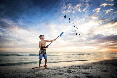 Full length of shirtless boy throwing sand in air with shovel at beach against cloudy sky during sunset