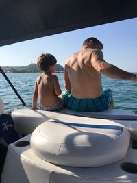 Shirtless father and son sitting in boat on sea against sky