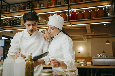 Female chef hand on shoulder of male chef writing in diary while standing in commercial kitchen