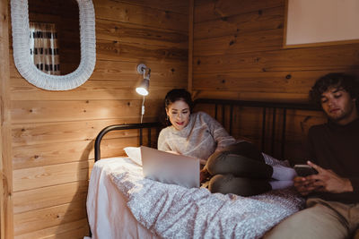 Young woman using laptop while sitting with male friend on bed at home