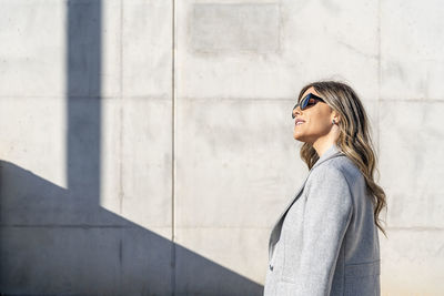Smiling businesswoman wearing sunglasses in front of wall on sunny day