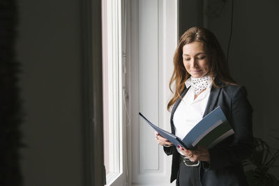 Mature female real estate agent reading brochure while standing by window at home