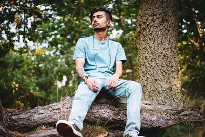 Full length of young man looking away while sitting on tree trunk