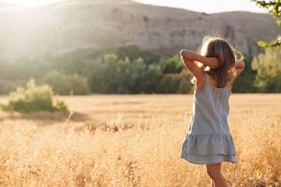 Rear view of girl with hand in hair standing on grassy field against mountain