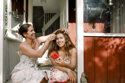 Smiling mother tying daughter's hair while sitting outdoors