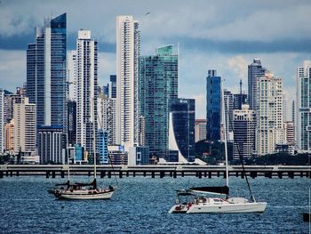 Sailboats in river by modern buildings against sky in city