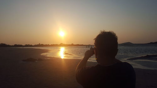 Rear view of silhouette man photographing at beach during sunset