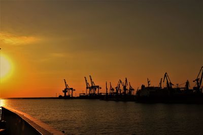 Silhouette cranes at harbor against sky during sunset