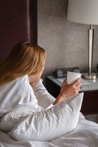 Woman holding coffee cup while lying on bed at hotel