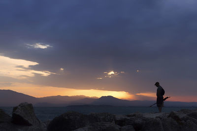 Man standing on rock at sea shore against sky during sunset