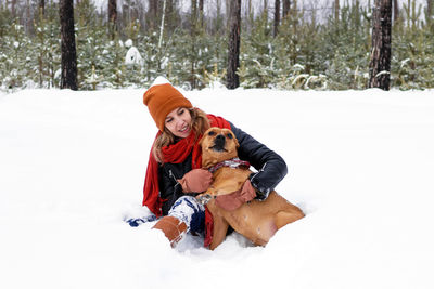 Beautiful young woman is sitting on a snow and playing with her dog in winter coniferous forest.