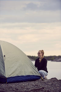 Smiling young woman crouching tent at beach against sky