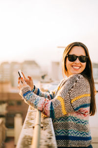 Portrait of smiling woman holding phone while standing by retaining wall