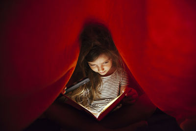 Close-up of girl with mobile phone light reading book under blanket