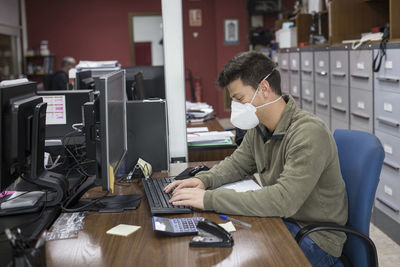 Side view of man wearing mask using computer on desk in office