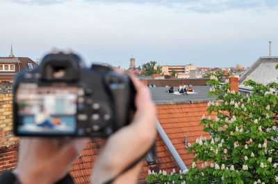 Cropped image of person photographing woman sitting on building terrace in city against sky