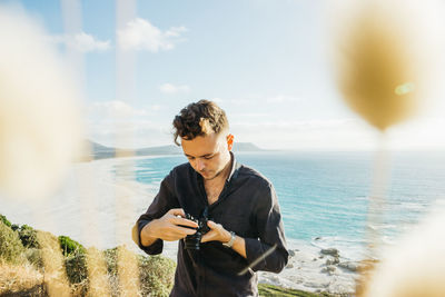 Young man using camera at sea against sky