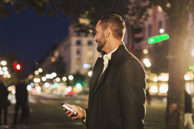 Side view of man looking away while holding smart phone on city street at night