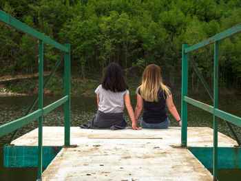 Rear view of women sitting on pier over water against trees