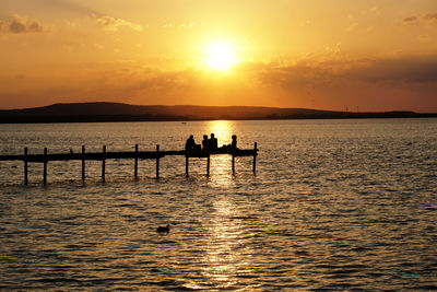 Silhouette people on pier in sea against orange sky