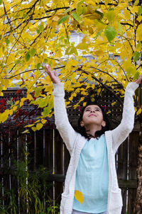 Smiling girl with arms raised touching with tree leaves during autumn