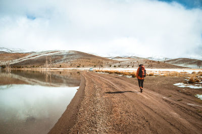 Rear view of man walking on dirt road against cloudy sky